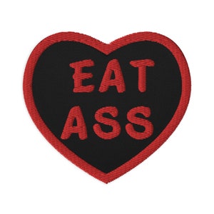 Może przedstawiać: Czerwona naszywka w kształcie serca z czarnego materiału z czerwonym haftem, na którym widnieje napis "EAT ASS".