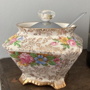 May include: A white porcelain sugar bowl with a floral pattern and gold accents. The bowl has a silver lid with a clear glass knob and a matching spoon.