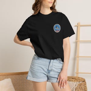 May include: Black t-shirt with a small, circular emblem on the left chest. The emblem features a detailed illustration of a ship, surrounded by text in a circular design. The shirt is paired with denim shorts.