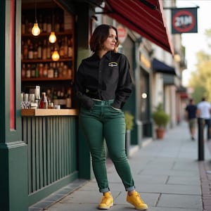 Puede incluir: Una persona con una camisa negra con un logo, jeans verdes y zapatillas amarillas está de pie frente a un bar. El bar tiene un toldo rojo y un letrero que dice "21M". El fondo incluye un bar con botellas y vasos.