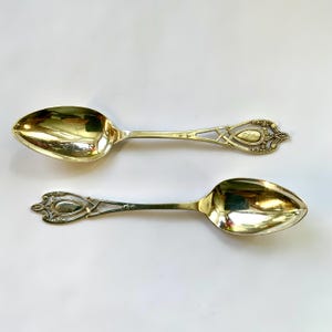 May include: Two antique silver spoons with ornate handles and oval bowls. The handles feature a decorative openwork design and an engraved detail. The spoons are displayed on a white surface, reflecting light.