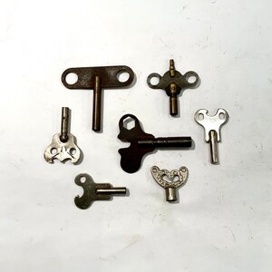 May include: A collection of seven antique skeleton keys with various designs and finishes, including brass, silver, and black. The keys are arranged on a white surface.