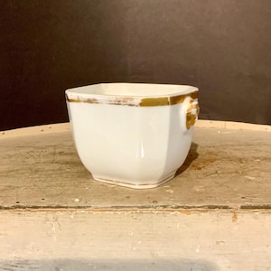 May include: A white ceramic square bowl featuring a gold-painted rim and a decorative gold-toned lion head relief on the side. The vessel has a smooth, glossy finish with subtle signs of wear on the metallic gold accents along the top edge.