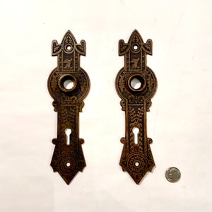 May include: Two antique brass door plates with intricate designs. Each plate features a keyhole and a circular opening for a knob. The plates have a dark patina and are in good condition.