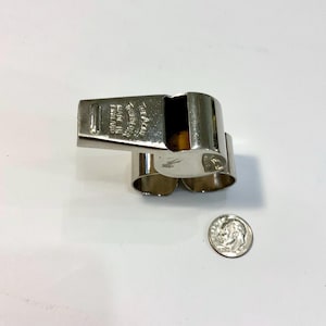 May include: A silver-colored whistle with a rectangular mouthpiece and two finger loops. The text "MADE IN ENGLAND" is visible on the mouthpiece. A U.S. dime is next to the whistle for size comparison. The whistle is likely used for sports or signaling.