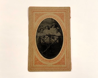 May include: An antique oval-shaped photograph of four people sitting on a bench. The photo is framed with a decorative border in shades of red and brown.