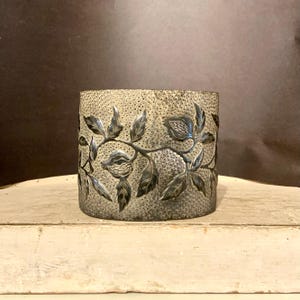 May include: A cylindrical, silver-toned container with a textured surface and a floral design. The design features detailed leaves and fruit, adding a touch of nature-inspired elegance to the piece. The container is likely used for storage or decoration.