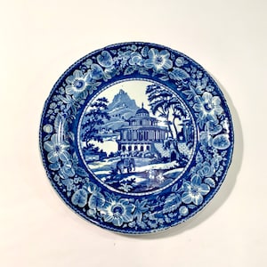 May include: A blue and white ceramic plate with a floral border and a central image of a building with a dome and a mountain in the background.