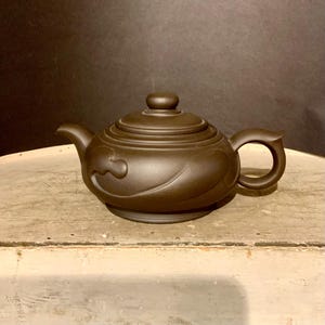 May include: A small, brown ceramic teapot with a curved spout and handle. The lid has a small knob and the body features a decorative swirl design. The teapot sits on a light-colored surface against a dark background.