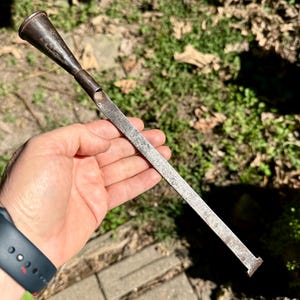 May include: A weathered, dark metal tool featuring a long, flat, rectangular shaft that terminates in a small, flared end. The opposite end of the tool transitions into a hollow, conical, funnel-shaped attachment with a dark, oxidized finish.