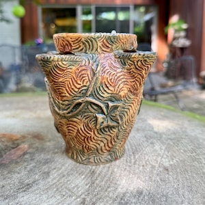 May include: A ceramic strawberry planter with a multi-tiered design featuring multiple openings. The surface has a textured, wavy pattern in shades of brown, tan, and muted green, with a raised relief of a leaf vine wrapping around the exterior.