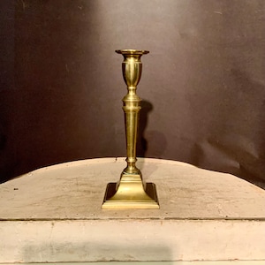 May include: A gold metal candlestick with a square base and a fluted column. The candlestick is on a light brown wooden surface.