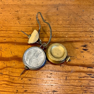 May include: Antique brass and silver-toned metal balance scale weights. The weights are round with a curved handle and a small wooden piece. The objects are on a wooden surface.