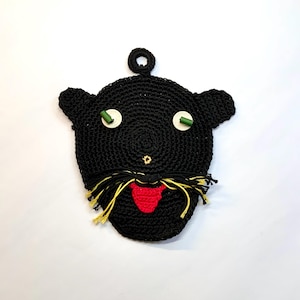 May include: A black crocheted cat-shaped potholder with a red tongue, yellow and black whiskers, and green and white eyes. The potholder has a loop for hanging and is made of thick yarn.