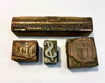 May include: A collection of vintage letterpress printing blocks. Includes a long block with the text "THE BOWMAN'S NATIONAL BANK" in reverse, an "EXIT" sign, a dollar sign, and a mechanical illustration.