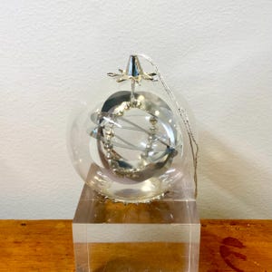 May include: A clear glass Christmas ornament with a silver metal interior design. The ornament has a silver metal cap and a silver string for hanging. The ornament is displayed on a clear acrylic block.