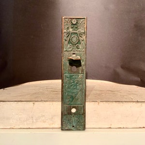 May include: A vintage, green-colored metal door plate with intricate floral and geometric designs. The plate has a small, rectangular handle and a round hole for a key.