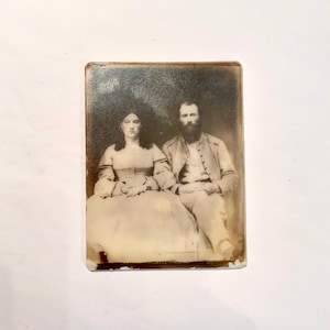 May include: A sepia-toned photograph of a seated couple. The woman wears a light-colored dress with long sleeves, and the man has a long beard and a jacket. The photograph has a slightly distressed appearance, with visible wear around the edges.