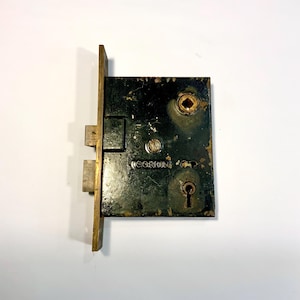 May include: Vintage black and gold door lock with the brand name "CORBIN" visible. The lock has a rectangular shape with a keyhole and a latch. The metal shows signs of wear, giving it a rustic appearance. The lock is likely made of metal.
