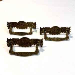 May include: Three antique brass drawer pulls with ornate detailing. Each pull features a decorative central panel with a detailed design and a curved handle. The brass has a warm, aged patina, suggesting vintage craftsmanship. These pulls are suitable for furniture restoration.