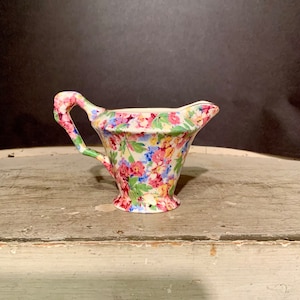 May include: A small, white creamer with a floral pattern in shades of pink, blue, yellow, and green. The creamer has a flared spout and a handle.