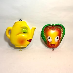May include: Two wall-mounted decorative plaques featuring anthropomorphic designs. One is a yellow teapot with closed eyes and red lips, and the other is a red and yellow apple with green leaves, wide eyes, and red lips. Each has a small metal hook.