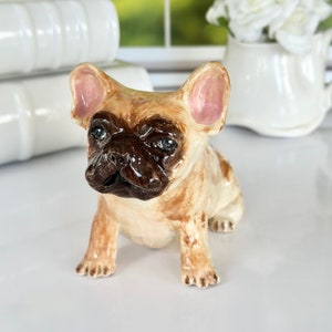 May include: A ceramic figurine of a fawn French bulldog with brown eyes and pink ears. The figurine is sitting on a white surface.