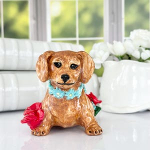 May include: A brown dachshund figurine with blue flower accents around its neck and red flowers on its side. The figurine is sitting on a white surface.