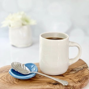 A hot cup of coffee has a blue heart shaped teaspoon rest next to it. The heart spoon rest has a silver spoon sitting on it.