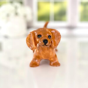 May include: A small, brown dachshund figurine with large, floppy ears and a black nose. The dog is standing, facing the viewer, with a glossy finish. The figurine is a decorative item, likely for a shelf or desk.
