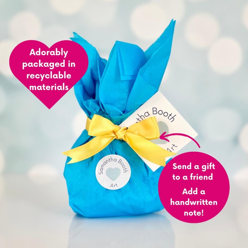 May include: A blue gift bag with a yellow ribbon and a white tag that says "Samantha Booth Art". The bag is wrapped in tissue paper and tied with a bow. The tag has a heart with the words "Samantha Booth Art" on it. The text "Send a gift to a friend Add a handwritten note!" is written on the tag.