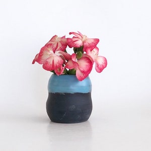 May include: A small, handmade ceramic vase with a two-tone design. The top half is a light blue, and the bottom half is a dark charcoal color. The vase holds a small bouquet of pink and white flowers, creating a simple, elegant floral arrangement.
