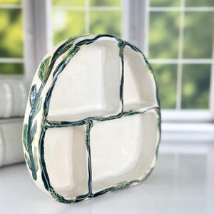 May include: A white ceramic divided serving dish with four compartments. The dish is decorated with green and dark blue leaf and vine designs around the edges. The dish is oval-shaped and sits on a white surface.