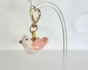 May include: A small ceramic bird charm featuring a glossy off-white body and soft pink wings. The bird is attached to a gold-toned metal lobster clasp by a small jump ring, allowing it to hang from a curved display stand for easy viewing.