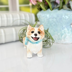 Puede incluir: Una figurita de cerámica de un perro corgi con una bufanda azul. La figurita es blanca con orejas marrones y una mancha marrón en la espalda. El perro sonríe y tiene una expresión feliz.