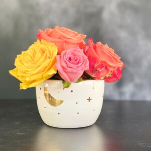 May include: A white ceramic planter with a gold crescent moon and stars design, filled with a bouquet of yellow, pink, and orange roses.