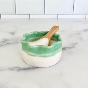May include: A white ceramic salt cellar with a green glaze and a wavy rim. A wooden spoon is inside the cellar, filled with white salt.