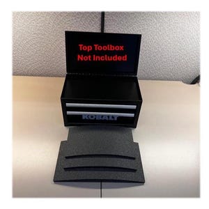 May include: A black Kobalt metal toolbox with two drawers and an open lid. The interior lid features red text that reads Top Toolbox Not Included. A separate black textured foam insert with three curved slots is positioned in front of the unit.