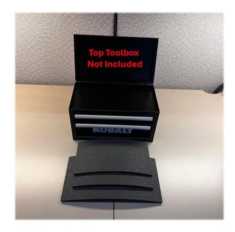May include: A black Kobalt metal toolbox with two drawers and an open lid. The interior lid features red text that reads Top Toolbox Not Included. A separate black textured foam insert with three curved slots is positioned in front of the unit.