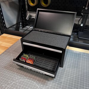May include: A black metal storage chest with a hinged lid and one pull-out drawer. The interior features black foam inserts with slots for organizing small items. Several small, rectangular spools of colored thread are stored in the drawer.