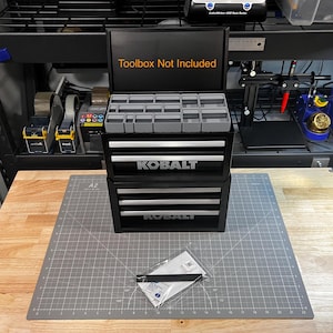 May include: A black three-drawer toolbox with the word "Kobalt" printed on the front of each drawer. The top drawer has a removable tray with multiple compartments. The toolbox is open and the text "Toolbox Not Included" is printed on the inside of the lid.