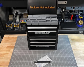 May include: A black three-drawer toolbox with the word "Kobalt" printed on the front of each drawer. The top drawer has a removable tray with multiple compartments. The toolbox is open and the text "Toolbox Not Included" is printed on the inside of the lid.