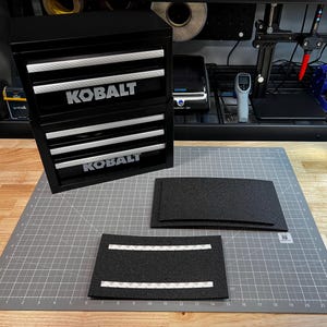 May include: A black Kobalt metal storage cabinet with two stacked units, each featuring three drawers with silver handles. Two rectangular black foam inserts with adhesive strips on the back are placed on a gray cutting mat with inch markings.