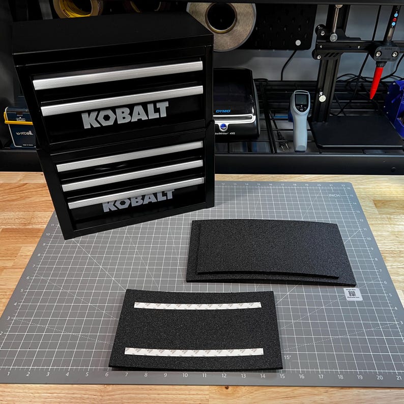 May include: A black Kobalt metal storage cabinet with two stacked units, each featuring three drawers with silver handles. Two rectangular black foam inserts with adhesive strips on the back are placed on a gray cutting mat with inch markings.