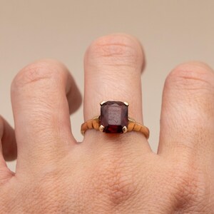 May include: A gold ring featuring a large, rectangular, dark red gemstone. The ring's band has a textured design. The gemstone is held in place by four prongs. The ring is on a finger.