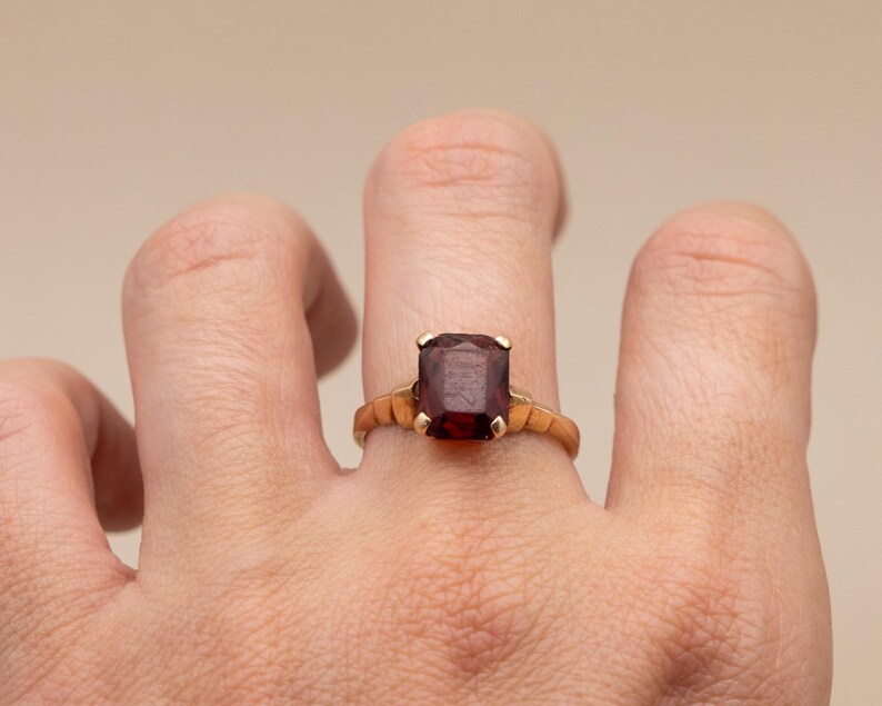 May include: A gold ring featuring a large, rectangular, dark red gemstone. The ring's band has a textured design. The gemstone is held in place by four prongs. The ring is on a finger.