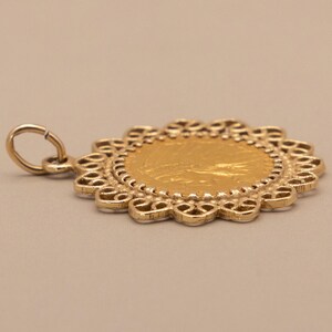 May include: A gold pendant with a circular design. The pendant features a central gold coin surrounded by a decorative, openwork border. A small loop is attached for hanging. The pendant is set against a neutral background.