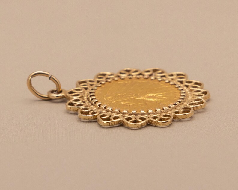 May include: A gold pendant with a circular design. The pendant features a central gold coin surrounded by a decorative, openwork border. A small loop is attached for hanging. The pendant is set against a neutral background.