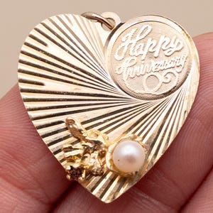 May include: A gold heart-shaped pendant featuring a sunburst pattern and a circular engraving that reads Happy Anniversary. The pendant is accented with a small white pearl and a tiny reddish gemstone set in a textured gold floral-like detail.