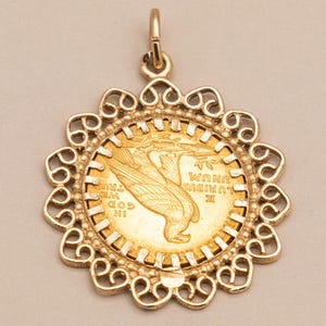May include: A gold pendant with a coin featuring an eagle design, set in a decorative filigree frame. The pendant has a loop for a chain and is shown against a neutral backdrop.